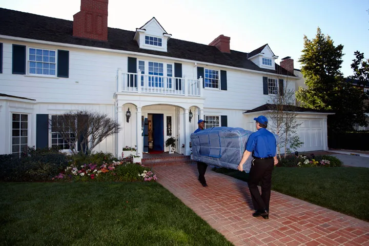 A mover carrying padded furniture out of a white colonial house